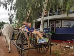 Erika Bud on an ox cart with the river cruise ship in the background along the Mekong River
